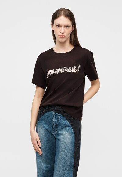 Νεαρός με μακριά μαλλιά που φοράει μαύρο μπλουζάκι με στάμπα και τζιν παντελόνι διχρωμίας μπλε και μαύρου, στέκεται μπροστά σε απλό ανοιχτό φόντο.