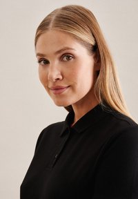 Czarna koszulka polo z kołnierzykiem, wykonana z fakturowanej tkaniny i z małym zapięciem na guziki. Widoczne gładkie włosy i naturalny makijaż.