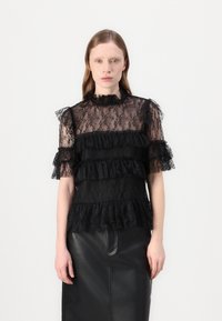 Top in pizzo nero con design a volants a strati, maniche corte e scollatura alta. Il tessuto ha una texture trasparente con motivi floreali intricati.