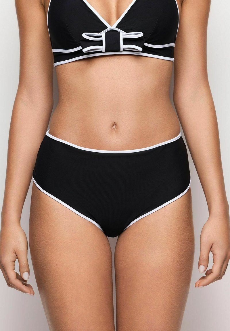 Kvinna klädd i svarta högmidjade bikiniunderkläder och matchande topp med vit kant och rosettdetalj, stående mot en neutral bakgrund.