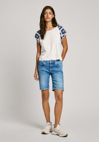 Biely krátky rukáv top s modrými vzorovanými detailmi, skombinovaný s svetlomodrými džínsovými šortkami. Model má na sebe béžové športové topánky.