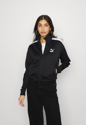 Chaqueta negra con cremallera hecha de una tela suave, con rayas laterales blancas y un logo de Puma. Combinada con pantalones negros y una parte superior blanca.