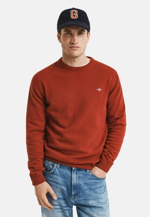 Pull en laine rouge avec un col rond et des poignets côtelés, associé à un jean bleu clair déchiré. La casquette présente une lettre "G" en orange.