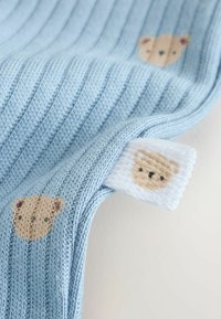 Tela de punto acanalada azul con pequeñas caras de oso marrón y una etiqueta blanca que también presenta una cara de oso tejida.