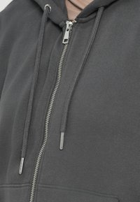 Siva zip-up dukserica s srebrnim patentnim zatvaračem i vezicama, izrađena od mekog materijala s glatkom teksturom i kengur džepom.