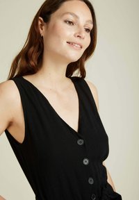 Robe noire sans manches avec un décolleté en V, cinq boutons noirs sur le devant et un tissu texturé.