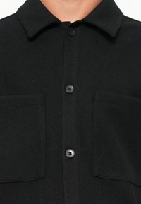 Black wool shirt with a collar, two chest pockets, and dark buttons. The fabric has a soft, textured finish.