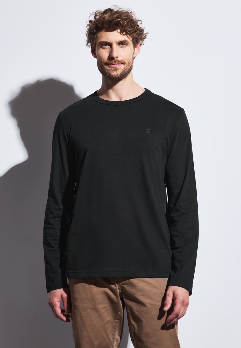 Czarna, długorękawowa koszula z gładkiego materiału, z okrągłym dekoltem, z dyskretnym logo na piersi, w zestawie z jasnobrązowymi spodniami.