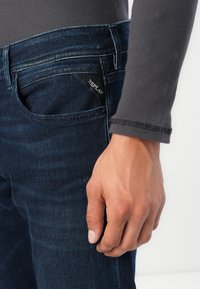 Jeans de mezclilla azul oscuro con un ajuste slim, que cuentan con un bolsillo cosido y una etiqueta de marca visible. La parte superior que lo acompaña es una camiseta de manga larga gris.
