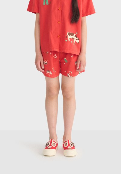 Bambino con maglietta rossa a maniche corte e pantaloncini abbinati con stampe di mucche e fiori verdi, scarpe slip-on rosse e bianche con dettagli floreali.