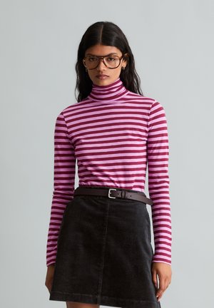 Frau mit großen braunen Brillen, pink-lila gestreiftem Rollkragenpullover und schwarzem Cordrock mit braunem Gürtel, stehend vor grauem Hintergrund.