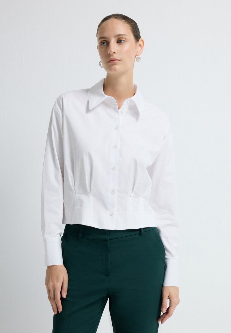 Witte, cropped button-up shirt met lange mouwen, klassieke kraag en een soepele textuur, gecombineerd met donkergroene, op maat gemaakte broek.