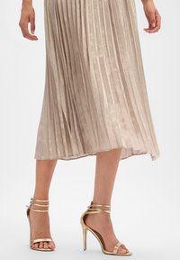 Femme portant une jupe midi plissée beige et des sandales à talons hauts dorées métalliques avec des brides à la cheville, posant devant un fond blanc.