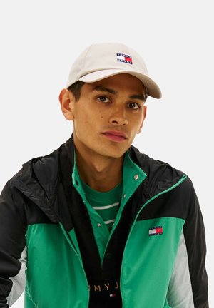 Joven con gorra beige de Tommy Jeans, camisa a rayas verde y chaqueta con bloques de color verde y negro con el logo de Tommy Jeans.