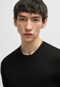 Black cotton crew neck sweater with a smooth texture, fitted design, and subtle stitching details around the neckline. Minimalist style.