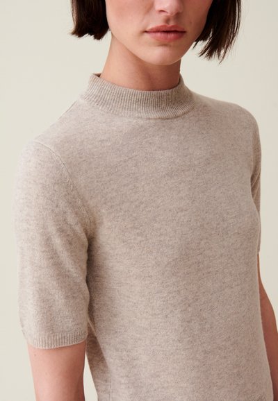 Maglione beige a maniche corte con colletto alto a coste. Texture morbida e design aderente, che mette in mostra una costruzione senza cuciture e senza hardware visibile.