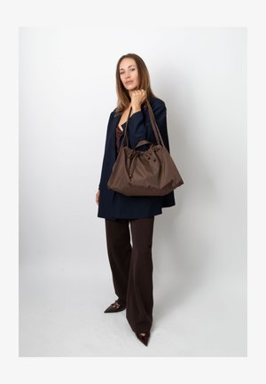 Ruskea nailonlaukku, jossa on kerätty yläosa, samanväriset olkahihnat ja sileä pinta. Malli pitää sitä yksiväristä valkoista taustaa vasten.