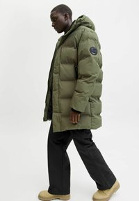 Man som går och bär en olivgrön dunjacka med huva, svarta byxor och beige stövlar mot en enfärgad vit bakgrund.