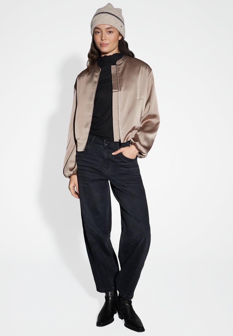 Satin bomber jacket in taupe, ribbed black top, wide-leg black jeans, and black heeled boots, with a light-colored knitted beanie.