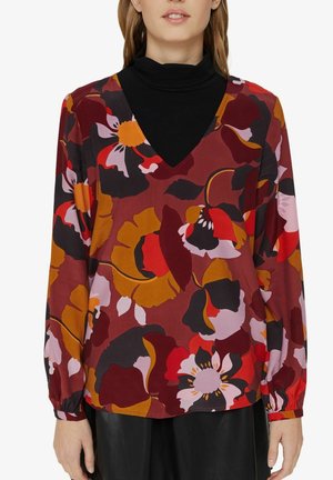 Frau trägt eine V-Ausschnitt-Bluse mit großen roten, orangefarbenen, schwarzen und pinken Blumenmustern über einem schwarzen Rollkragenpullover.