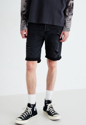 Shorts in denim nero con dettagli invecchiati, orli arrotolati e vestibilità slim. Indossati con sneakers alte nere e calze bianche.