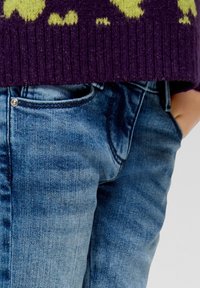 Maglione lavorato a maglia viola con design giallo, abbinato a jeans di denim blu. I jeans presentano un dettaglio sulla tasca e una texture leggermente consumata.