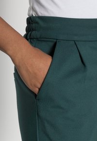 Gröna tygbyxor med sidofickor, med en veckad design och resårmidja. Slät textur och skräddarsydd passform är synliga.