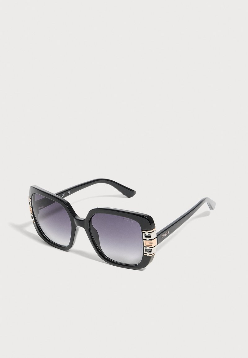 Lunettes de soleil noires à monture carrée avec verres foncés dégradés et détails décoratifs dorés sur les charnières, présentées sur un fond uni.