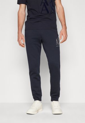 Mann trägt schwarze Armani Exchange Jogginghose und passendes T-Shirt mit weißen Sneakern und steht vor einem schlichten Hintergrund.