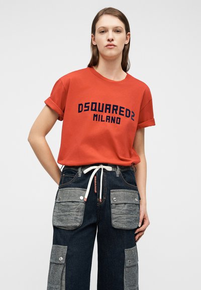 Γυναίκα που φοράει κόκκινο T-shirt με γυρισμένα μανίκια και το κείμενο "DSQUARED2 MILANO" και σκούρο παντελόνι με μεγάλες γκρι τσέπες cargo και λευκή ζώνη με κορδόνι.