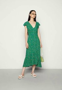 Mulher em vestido midi floral verde, sandálias brancas, pequena bolsa verde e óculos de sol com tonalidade verde, contra um fundo claro e simples.