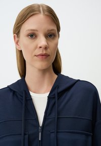 Sweat à capuche zippé en coton navy avec cordon de serrage, coutures horizontales et poches avant, superposé sur une chemise blanche.