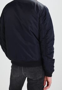 Giacca bomber blu navy con una texture liscia e imbottita, polsini e orlo a costine, e un design semplice e privo di ornamenti.