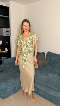 Femme portant un chemisier à imprimé feuilles vertes et une jupe beige, debout près d'un canapé bleu dans le salon, tenant un petit sac beige.