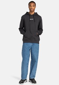 Schwarzer Kapuzenpullover mit Fronttasche, weißem Logo, kombiniert mit blauen Jeans und schwarzen Sneakers. Lässiges, locker sitzendes Design.