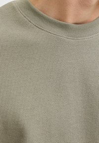 Lähivõte inimesest, kes kannab heledat oliivirohelist vahvlitekstiilist kampsunit, millel on sooniline ümarlõikega krae.