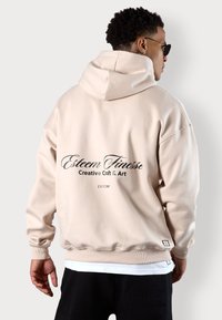Beige übergroßer Hoodie mit Kapuze und Kordelzug, gerippten Bündchen und Saum. Auf der Rückseite befindet sich schwarzer Drucktext: "Esteem Finesse Creative Craft & Art."
