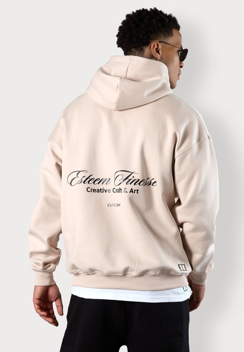 Beige übergroßer Hoodie mit Kapuze und Kordelzug, gerippten Bündchen und Saum. Auf der Rückseite befindet sich schwarzer Drucktext: "Esteem Finesse Creative Craft & Art."