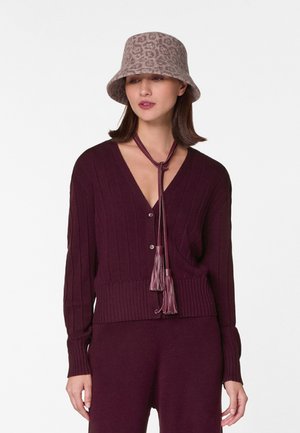 Cardigan en tricot bordeaux avec texture côtelée et décolleté en V. Se ferme avec des franges au col, associé à un chapeau seau à motifs.