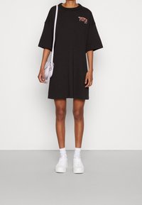 Czarna, oversize'owa sukienka T-shirt z okrągłym dekoltem i czerwonym logo "PUMA", zestawiona z białymi sneakersami i małą lawendową torebką.