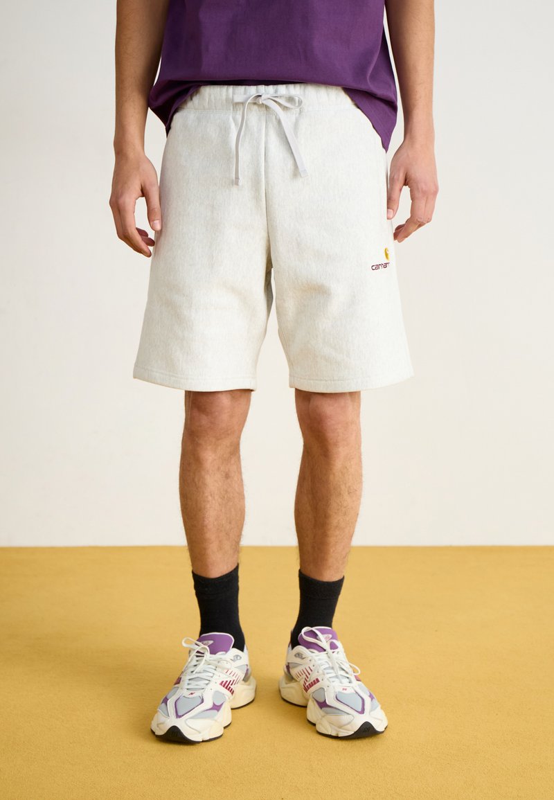 Pantalones cortos de sudadera grises con cordón, que presentan un logo bordado en la pierna. Combinados con zapatillas blancas y moradas.