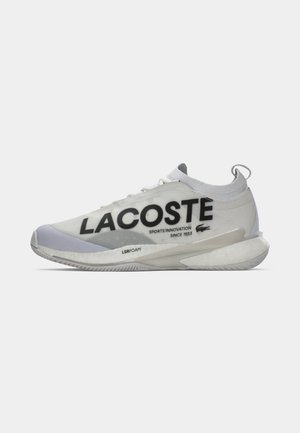 Sneaker Lacoste bianca e grigia con colletto simile a una calza, grande logo nero con testo, e suola imbottita etichettata "LSRFOAM" su uno sfondo bianco.