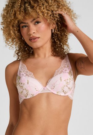 Soutien-gorge en dentelle rose avec motif floral, bonnets souples, bords festonnés et un bouton décoratif au centre. Légèrement transparent avec bretelles réglables.