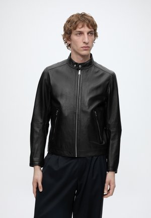 Jonge man met een zwarte leren jas met rits en opstaande kraag en donkere broek, staand tegen een effen lichte achtergrond.