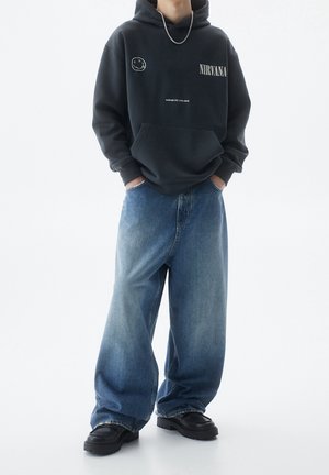 Persoon draagt een zwarte Nirvana-hoodie met een smiley-gezicht grafiek, losse blauwe spijkerbroek, zwarte dikke schoenen en een zilveren ketting, handen in de zakken.