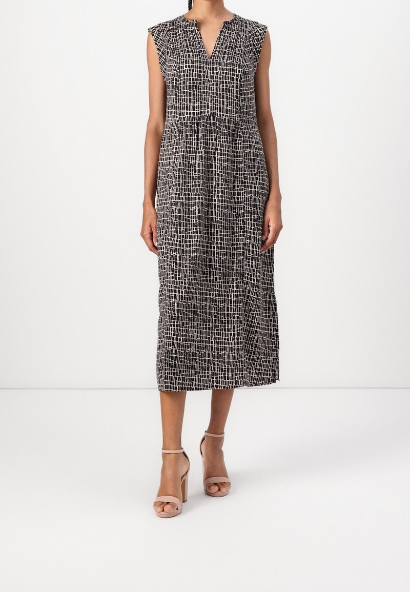 Femme portant une robe midi sans manches à motif noir et blanc avec un décolleté en V et des sandales beige à talons hauts, debout devant un fond uni.