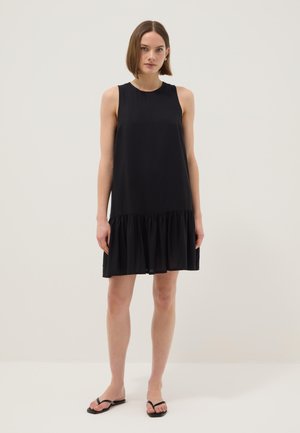 Frau steht in einem ärmellosen schwarzen Kleid mit gerafftem Saum und schwarzen Flip-Flop-Sandalen vor einem schlichten hellen Hintergrund.