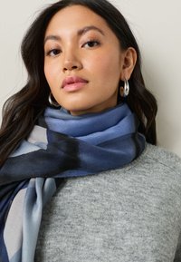 Blue and gray patterned scarf made from soft fabric, draped around the neck, complemented by a light gray textured sweater.