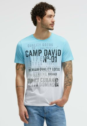 Mann mit lockigem Haar, der ein blau-weißes grafisches T-Shirt und blaue Jeans trägt, steht mit einer Hand in der Tasche und schaut nach rechts.