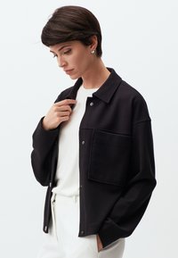 Zwarte geweven jas met een kraag, voorzak en drukknoopsluiting, gedragen over een witte shirt, gecombineerd met lichte broek.